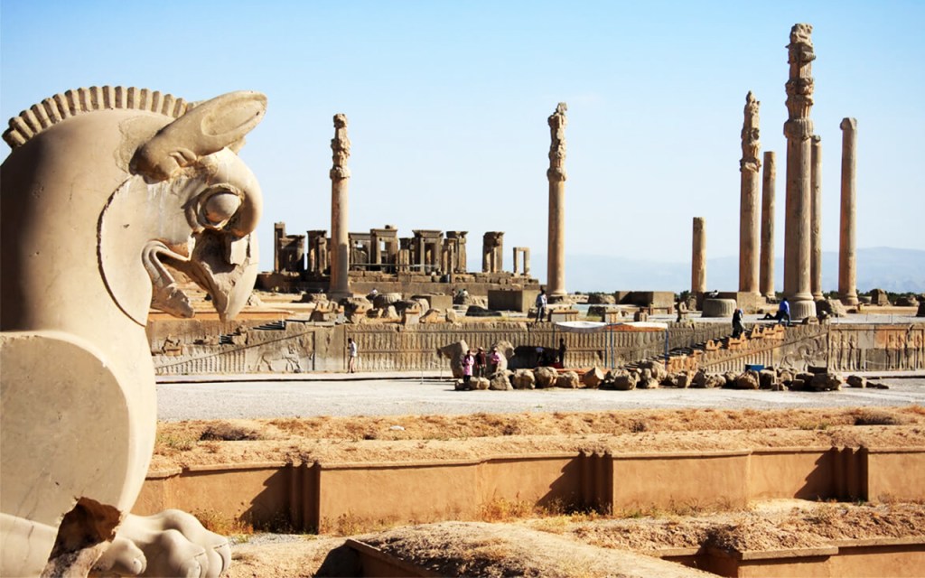 A close-up of a carved stone griffin sculpture in the foreground, with ancient columns and ruins of Persepolis in the background under a clear blue sky.